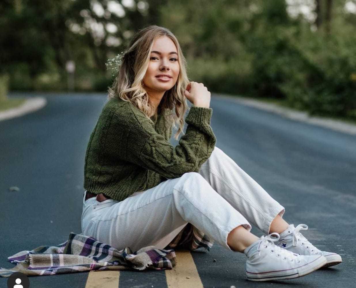 Young woman sitting on road with a plaid blanket, wearing a green sweater and white sneakers.