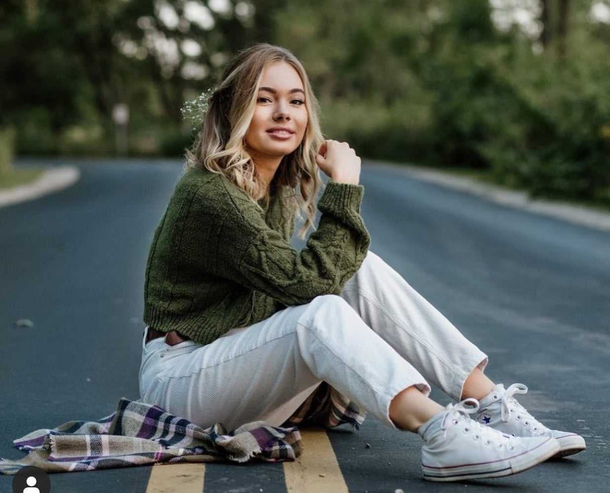 Young woman sitting on road with a plaid blanket, wearing a green sweater and white sneakers.