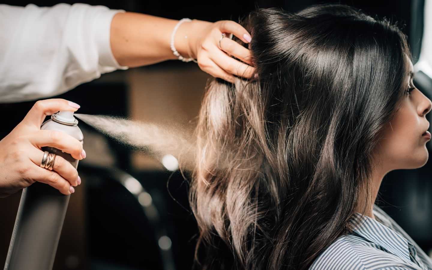Hair stylist applying hairspray to woman's styled dark hair.