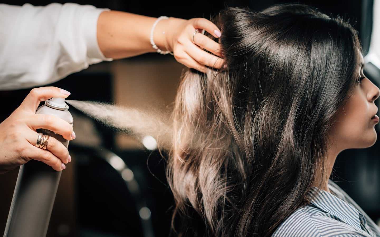 Hair stylist applying hairspray to woman's styled dark hair.