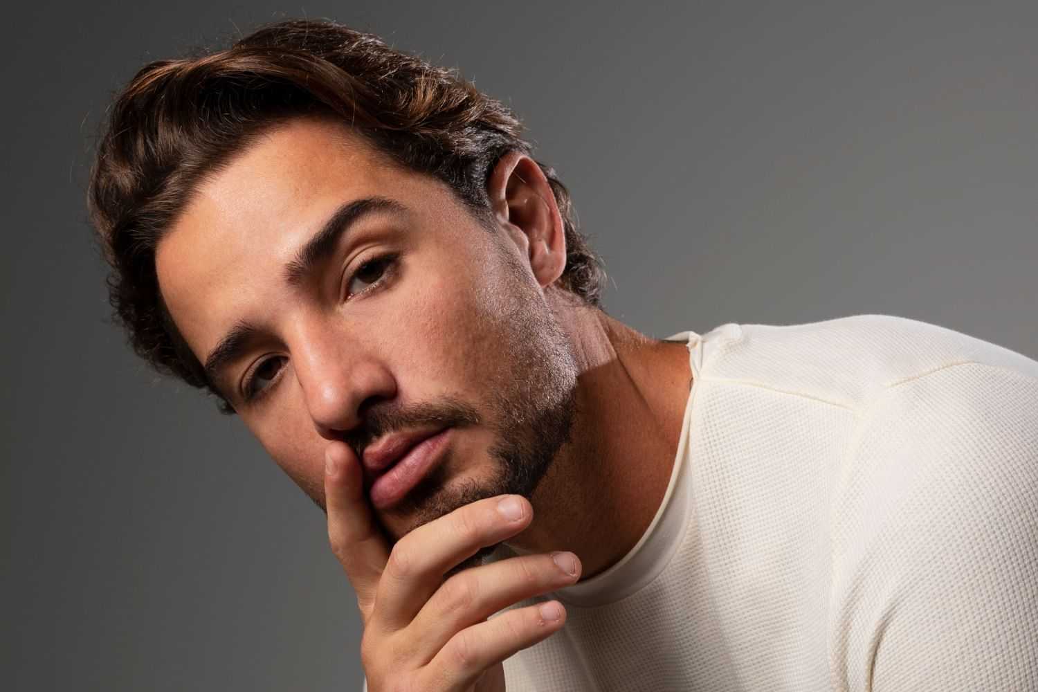 Man in white shirt with thoughtful expression, hand on chin against gray background.